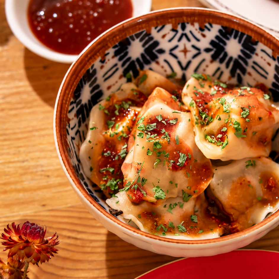 A close-up of dumplings in a patterned ceramic bowl with a spicy sauce and herb garnish, set on a wooden table with other dishes partially visible.